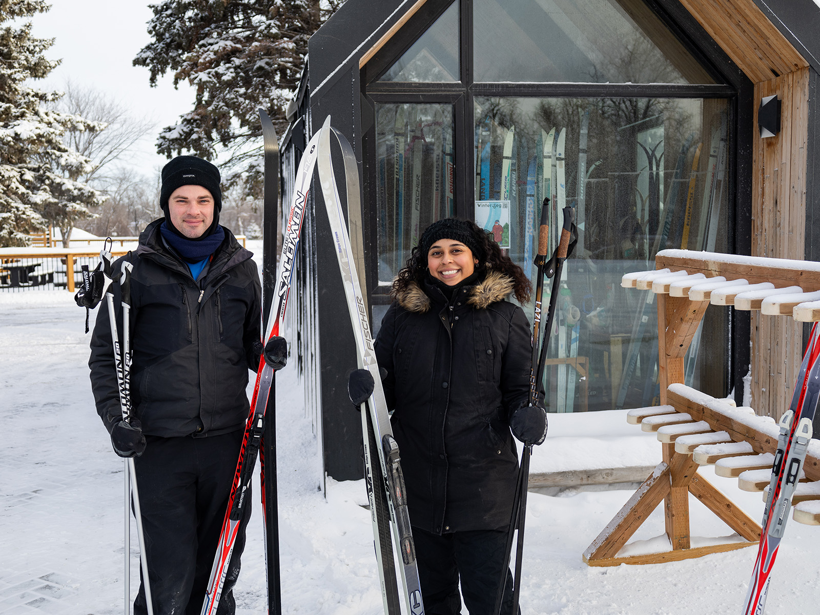 a man and woman holding skiis stand in front of a rental shack